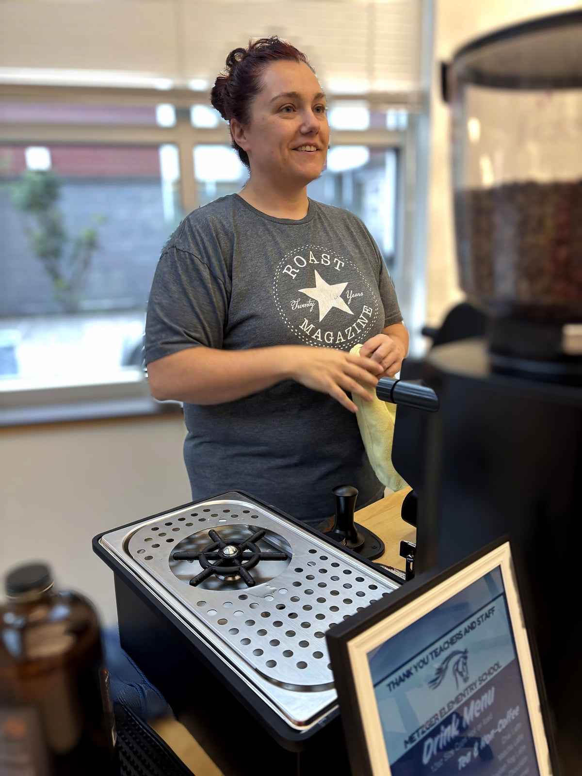 A woman wearing a gray t-shirt with a white 'Roast Magazine' logo stands behind a coffee preparation station inside a cafe. A coffee grinder filled with coffee beans is visible on the right, and a metal drip tray sits in the foreground.